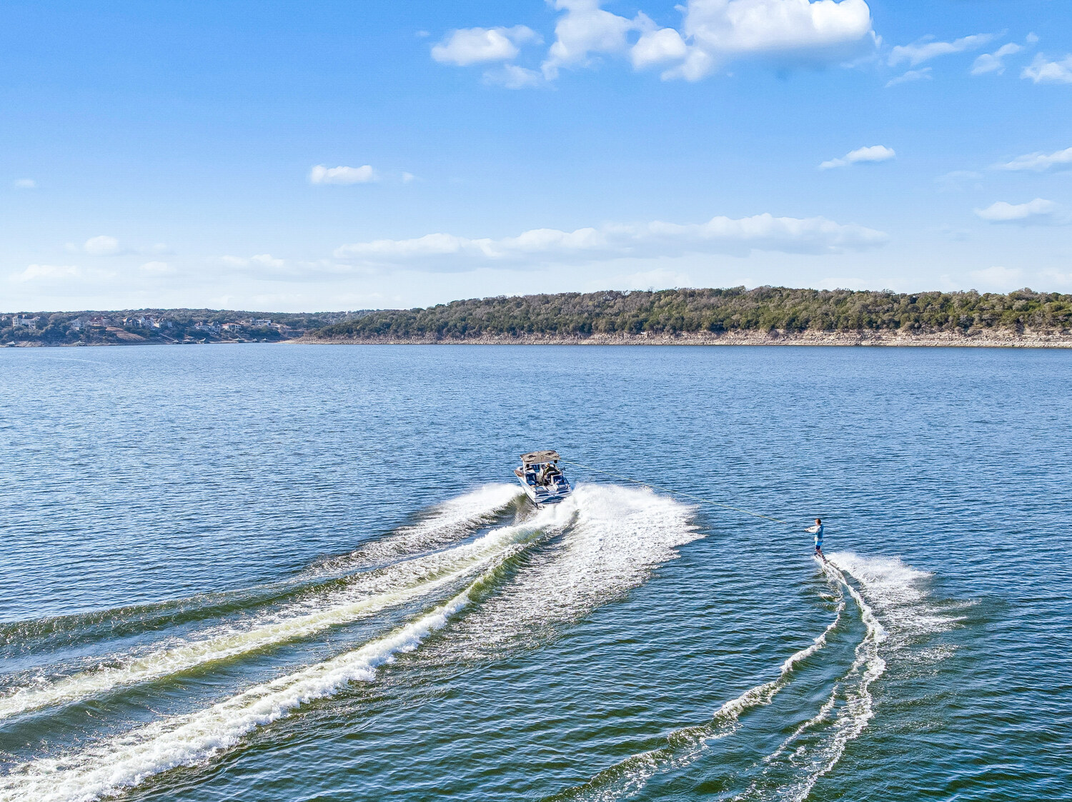 Water skier behind a boat on open water on Lake Travis, with the wooded shoreline and lake houses of the Hill Country in the background